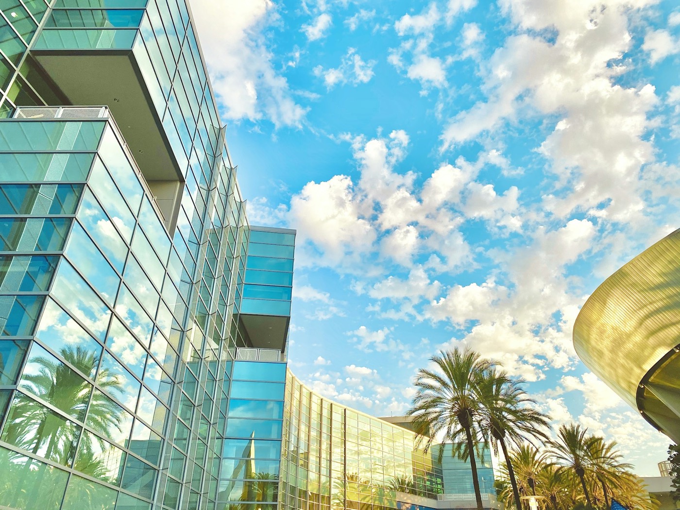 Glass towers of the Anaheim Convention Center with palm trees under a cloudy blue sky