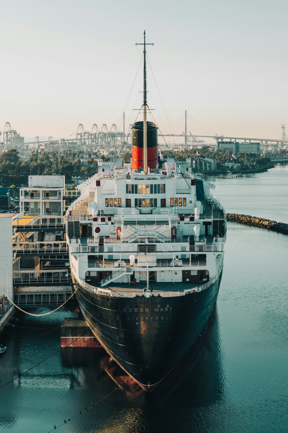 The Queen Mary ocean liner docked in Long Beach with port cranes and a bridge behind