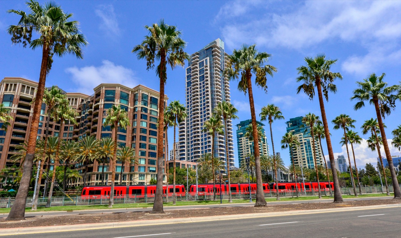 A red San Diego trolley passing palm trees and downtown towers